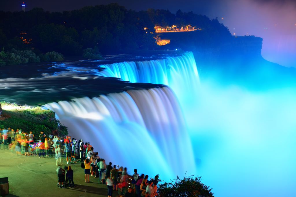 Visitors at a railing watching the American Falls lit in blue at night in Niagara Falls State Park