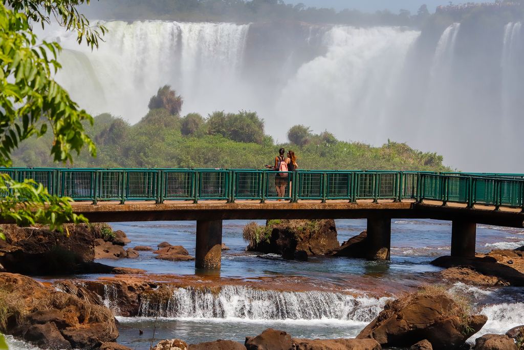 Visitors on a green walkway over the river with Iguazu Falls behind them