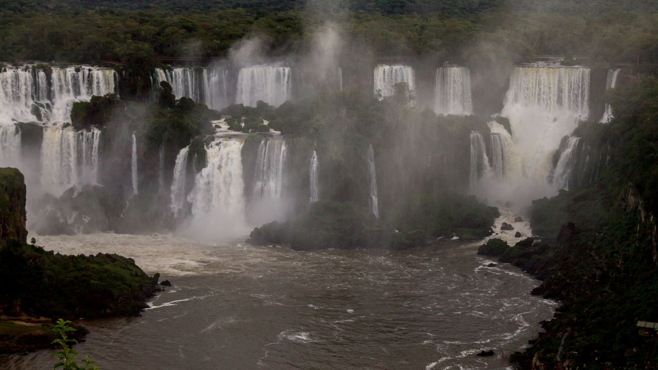 Visitors on a walkway at Iguazu Falls with waterfalls on both sides under a cloudy blue sky