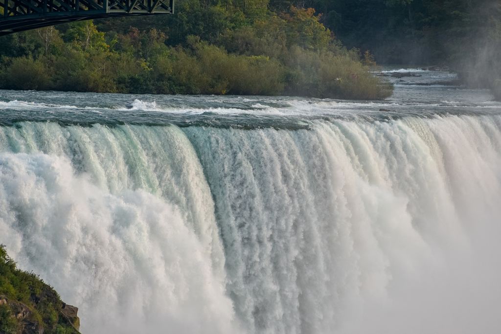 A thick sheet of white water pouring over the crest of Niagara Falls with a bridge above on the left