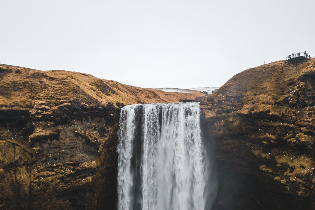 A tall waterfall dropping over a cliff between dry, brown hillsides with a few visitors on the rim