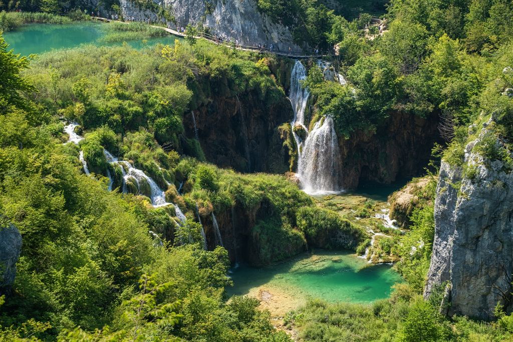 A waterfall dropping into a turquoise lake with a footpath along the cliff above in Plitvice Lakes National Park