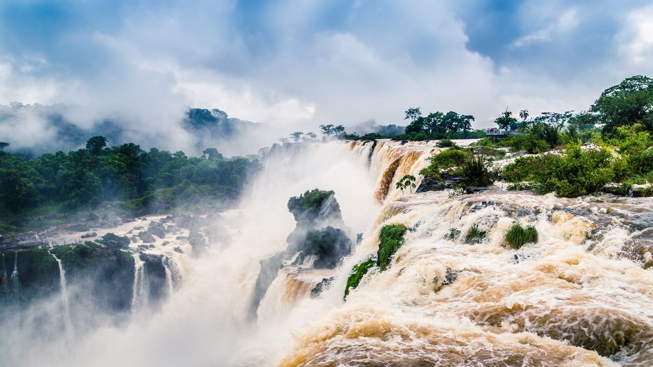A wide waterfall with brown water and drifting mist above a forested gorge under low cloud