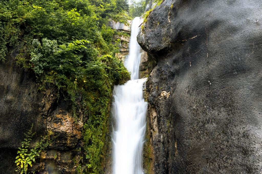 A waterfall dropping in two stages between dark rock walls and green plants