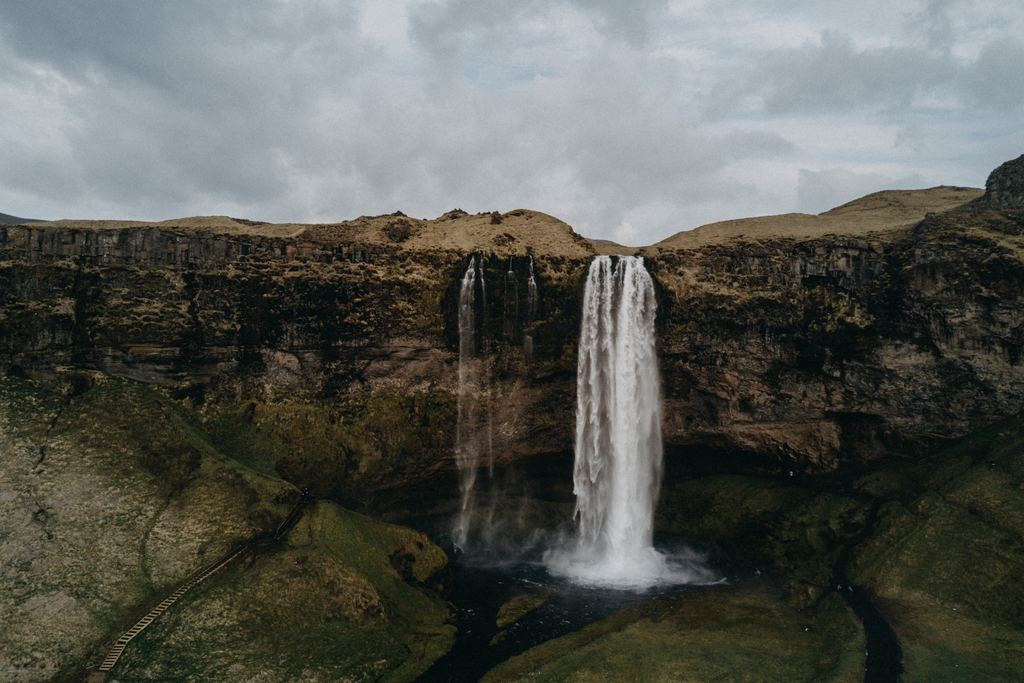 A tall waterfall dropping over a dark cliff into a pool, with a path along the slope and a cloudy sky