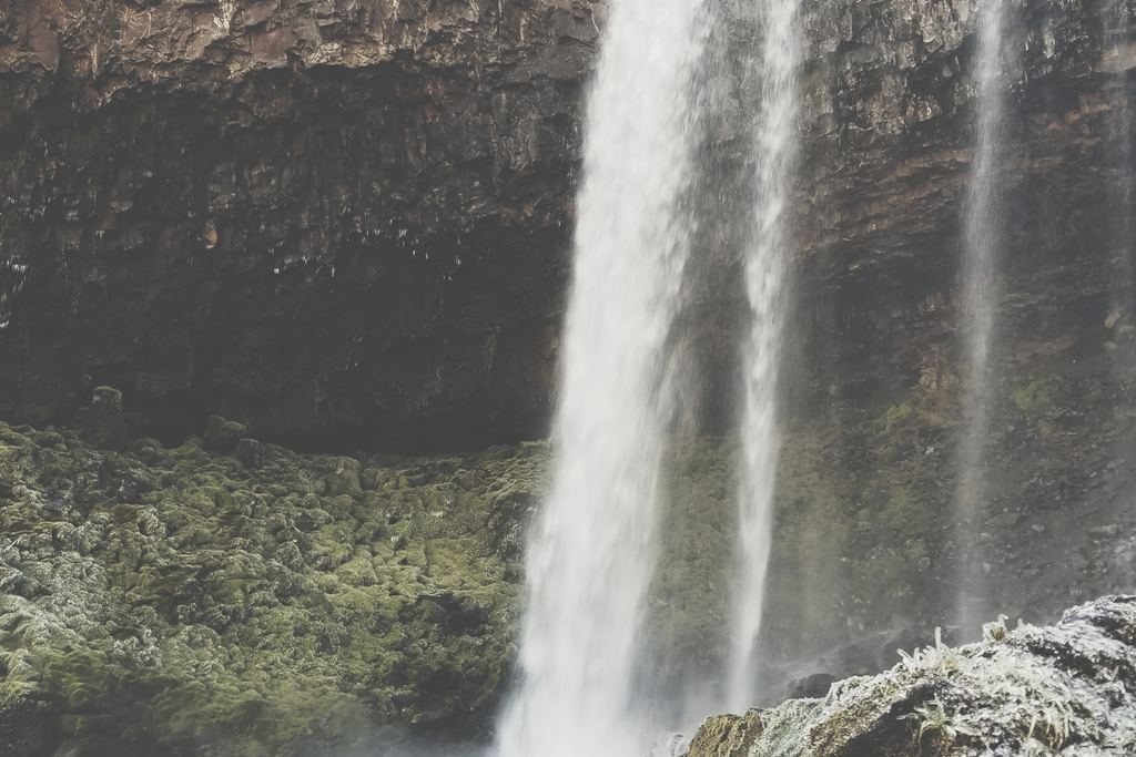 A tall waterfall dropping over a dark rock wall into a shallow pool with mossy rocks