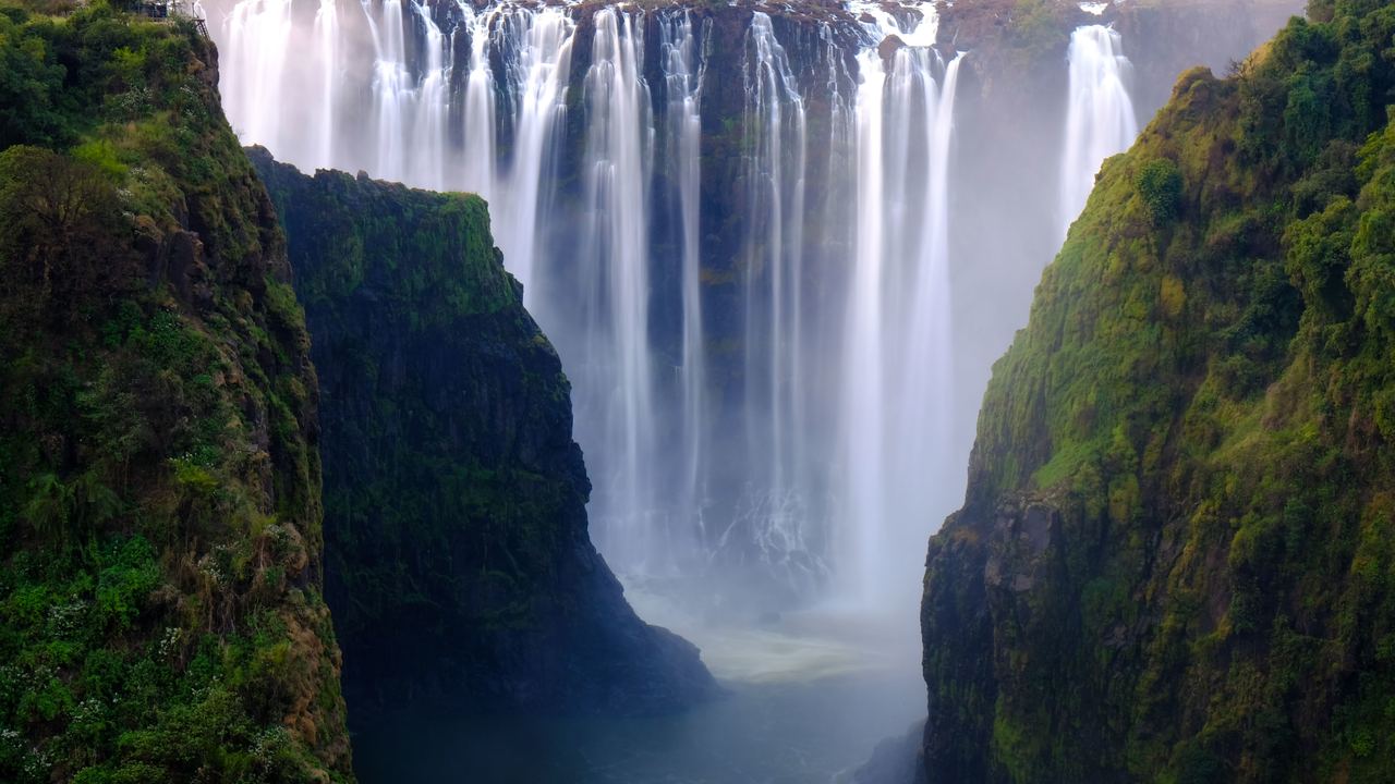 A wide curtain of waterfalls dropping into a deep green gorge with mist at its base