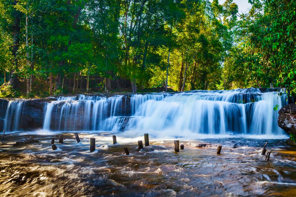 Wide, low waterfall in Phnom Kulen National Park flowing over a rock ledge between trees