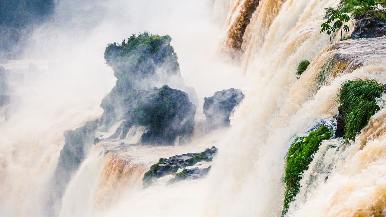 A wide waterfall in flood, brown water pouring over a rocky edge under a cloudy sky with spray rising