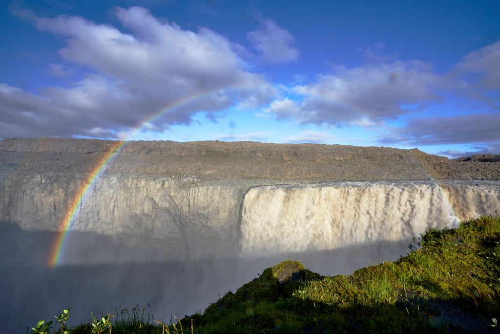 A wide waterfall with a rainbow seen from the grassy rim in northeast Iceland